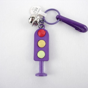 May include: A purple traffic light keyring with red, yellow, and green lights. The keyring has a silver bell and a purple clasp. A small tag with text is attached to the keyring.