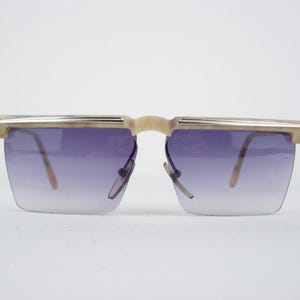 May include: Rimless sunglasses with a gradient purple tint. The frames are a light beige color with a silver accent across the top. The lenses are square-shaped and the arms are thin and light brown.