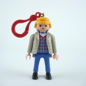 Puede incluir: Una figura de Playmobil de una persona con una chaqueta beige, pantalones azules y zapatos negros. La figura tiene un sombrero amarillo y un clip rojo sujeto a la cabeza. La camisa tiene un estampado de cuadros azules y rojos.