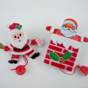 May include: Two Christmas-themed ornaments. One is a Santa Claus figure with a red and white suit, holding a bell and a sign that reads "Merry Christmas." The other is Santa Claus emerging from a red brick chimney with a bell attached.
