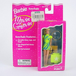 Può includere: Una bambola portachiavi Barbie Movin' Groovin' in una scatola rosa. La bambola ha i capelli biondi e indossa un vestito verde neon e blu. La scatola presenta il logo Barbie e il testo "Keychain Features".