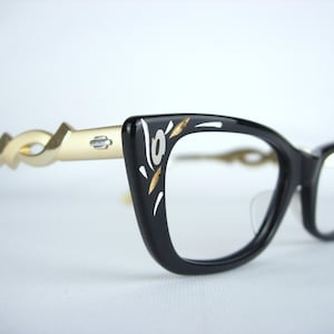 May include: Black cat-eye glasses with gold-colored accents. The frames are black with white and gold decorative elements. The temples are a light gold color with a unique, swirling design.
