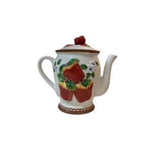 Könnte beinhalten: Eine weiße Keramik-Teekanne mit einem erhabenen, dreidimensionalen Design aus roten Äpfeln, grünen Blättern und kleinen weißen Blüten. Der Deckel hat einen roten Apfel als Knauf, und der Boden sowie der Deckelrand sind braun strukturiert.
