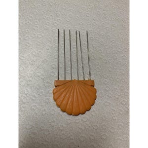 Könnte beinhalten: Ein Haarkamm mit einem pfirsichfarbenen Kunststoffgriff in Form einer Muschel, von dessen Oberkante sechs dünne, vertikale Metallzinken ausgehen. Der Griff weist ein strukturiertes, fächerartiges Design mit strahlenförmigen Rillen auf.