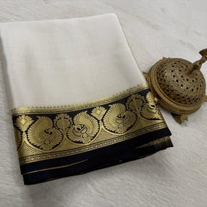 May include: A folded off-white fabric textile featuring a wide black border adorned with intricate gold metallic peacock and floral motifs, accompanied by a small, ornate, dome-shaped brass incense burner with detailed cut-out patterns.