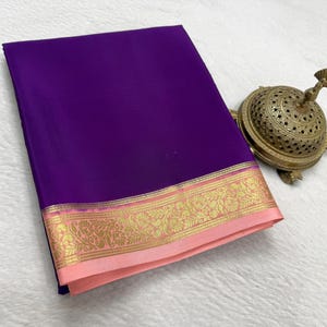 May include: A folded deep purple fabric textile featuring a wide border with a light pink base and intricate gold metallic floral patterns. Beside the fabric sits a small, round, ornate brass incense burner with a perforated dome lid.