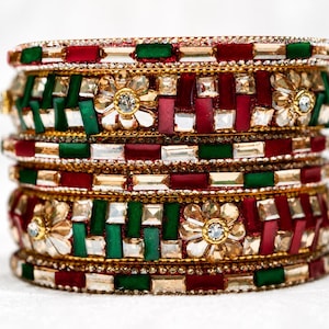 May include: A stack of gold-toned bangles featuring alternating rectangular red and green stones, clear faceted crystals, and floral-shaped accents with central clear stones. The bangles are arranged in a vertical column with a metallic finish.
