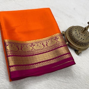 May include: A vibrant orange fabric folded neatly, featuring a wide magenta border adorned with intricate gold-toned peacock motifs and geometric patterns, accompanied by a metallic, dome-shaped incense burner with detailed filigree cutouts.