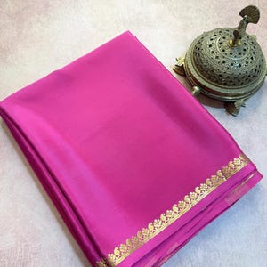 May include: A vibrant fuchsia pink fabric folded neatly, featuring a metallic gold border with a repeating floral and paisley pattern along the edge. Beside the fabric sits a small, ornate, dome-shaped metallic incense burner with intricate cutouts.