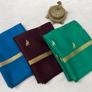May include: Three folded fabric pieces in blue, deep maroon, and green, each featuring a gold metallic border and small gold bird motifs. A small, round, ornate gold-toned metal container with a perforated lid sits above the fabric pieces.