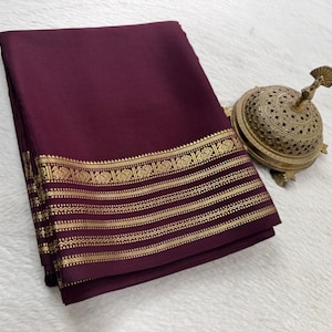 May include: A folded deep burgundy fabric with a wide gold border featuring intricate repeating peacock motifs and horizontal geometric lines, accompanied by a small, ornate, dome-shaped metallic gold incense burner with cut-out patterns.