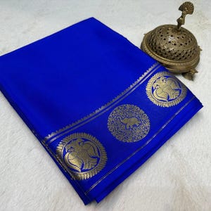May include: A folded vibrant royal blue fabric featuring a wide border with intricate gold metallic circular motifs depicting elephants and birds, accompanied by a small, ornate, perforated brass incense burner with a peacock finial.