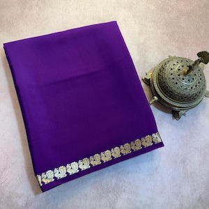May include: A folded deep purple fabric textile featuring a horizontal border pattern of repeating gold-colored peacock motifs along the bottom edge, positioned next to a metallic, dome-shaped incense burner with intricate cut-out details.