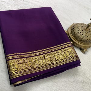 May include: A folded deep purple fabric textile featuring a wide, ornate gold metallic border with intricate woven floral and paisley patterns, accompanied by a small, round, perforated brass incense burner with a decorative finial top.
