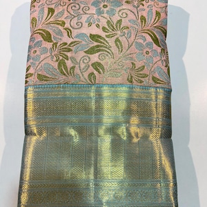 May include: A folded textile featuring a light pink background with a woven floral pattern in light blue and olive green. The lower section displays a wide, metallic gold border with intricate geometric detailing and a solid light blue edge.