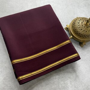 May include: A folded deep burgundy fabric textile featuring two parallel horizontal gold-colored bands with a repeating diamond-shaped pattern along the edge. Beside the fabric sits a round, ornate, metallic gold-toned incense burner.