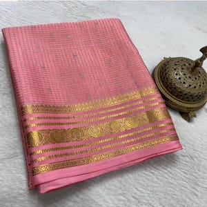 May include: A folded pink fabric with a subtle grid pattern and small silver accents, featuring a wide, ornate gold metallic border with intricate circular and geometric motifs, positioned next to a decorative, perforated brass incense burner.