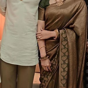 May include: A light sage green long-sleeved tunic with a deep V-neckline paired with olive green trousers, and a dark bronze-toned saree featuring a subtle woven pattern, a dark green border with gold paisley motifs, and gold jewelry including a necklace.