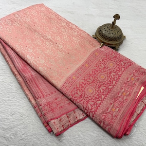 May include: A folded peach-colored textile featuring an intricate woven floral pattern in a lighter shade, complemented by a wide, deep pink border with detailed circular motifs and a small, ornate, metallic, dome-shaped incense burner.