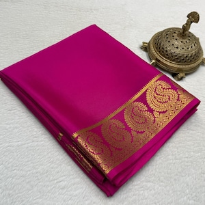 May include: A folded magenta fabric textile featuring a wide border with a gold-colored metallic paisley pattern. Beside the fabric sits a small, round, ornate brass-colored metal container with a perforated lid and a decorative finial top.