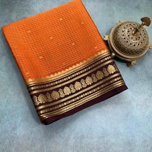 May include: A folded orange fabric with a gold grid pattern and a wide dark purple border featuring intricate gold floral motifs, accompanied by a metallic, dome-shaped incense burner with detailed filigree and a pointed finial top.