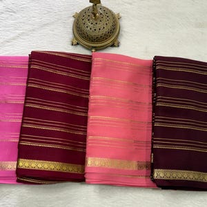 May include: Four folded fabric pieces in shades of bright pink, deep burgundy, light coral, and dark plum. Each piece features horizontal gold-toned metallic stripes and a decorative gold-toned border along the bottom edge of the fabric.