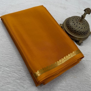 May include: A folded mustard yellow fabric with a smooth texture and a metallic gold border featuring a repeating peacock motif. Beside the fabric sits a round, perforated brass incense burner with a decorative finial on its lid.