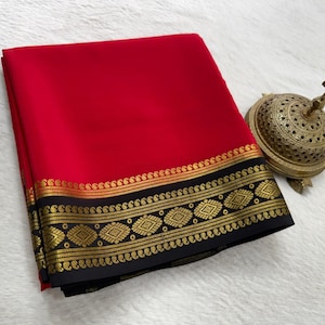 May include: A folded red saree featuring a wide black border adorned with intricate gold metallic patterns, including paisley motifs and geometric shapes, accompanied by a small, ornate, dome-shaped brass incense burner.