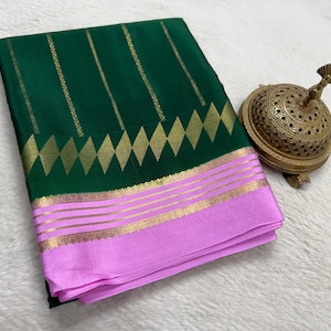May include: A folded dark green saree featuring vertical gold stripes and a row of gold diamond shapes above a light pink border. The border is decorated with horizontal gold lines and intricate gold patterns along the edges.