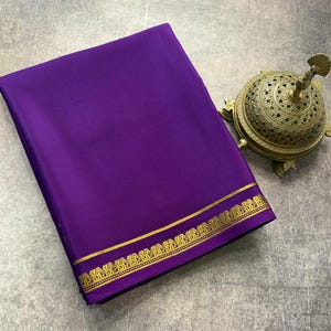 May include: A folded deep purple fabric textile featuring a horizontal gold border with a repeating elephant motif. Beside the fabric sits a round, ornate, metallic gold incense burner with a perforated dome lid and three small feet.