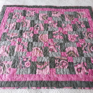 May include: A square patchwork quilt featuring a repeating checkerboard pattern of alternating dark gray textured fabric squares and pink floral patterned squares, finished with a wide pink border and a thin dark gray outer edge trim.