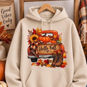 Op de afbeelding: Een beige hoodie met een grafische print van een oranje pick-up truck gevuld met pompoenen, zonnebloemen en herfstbladeren. De laadbak toont de tekst Hello Fall met een hartje, vergezeld door een koffiebeker en laarzen.