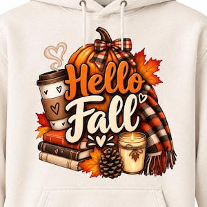 May include: A cream-colored hooded sweatshirt featuring a graphic print of a pumpkin, a coffee cup, a stack of books, a candle, a pinecone, and autumn leaves. The text "Hello Fall" is printed in the center with a plaid scarf accenting the design.