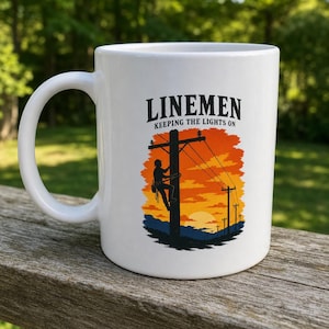 Peut inclure: Un mug en céramique blanche orné d'une illustration graphique représentant un technicien sur un poteau électrique devant un coucher de soleil orange et jaune. Le texte au-dessus indique LINEMEN KEEPING THE LIGHTS ON en lettres noires.