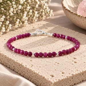 May include: A delicate bracelet featuring small, faceted, deep pink ruby-coloured beads interspersed with tiny, smooth, polished silver-toned metal spacer beads. The bracelet is secured with a silver-toned lobster claw clasp and a small jump ring.