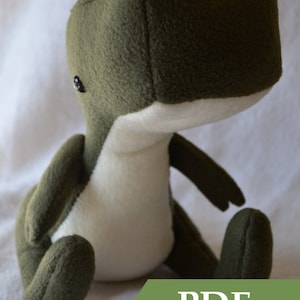 May include: A plush green and white dinosaur toy sitting on a white surface. The toy has a large head and small arms and legs. The text "PDF PATTERN" is visible in the lower right corner of the image.