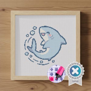 May include: A cross-stitch embroidery of a light blue shark with a white belly, pink cheeks, and a smiling mouth, surrounded by small blue bubbles. The artwork is displayed in a square light-colored wooden frame with a visible grain texture.