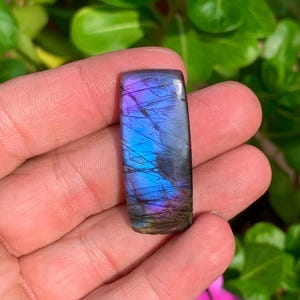 May include: A rectangular labradorite gemstone cabochon featuring a polished surface with iridescent flashes of vibrant blue and purple hues, contrasted by dark grey inclusions and fine natural surface fractures throughout the stone.