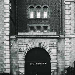 Puede incluir: Una foto en blanco y negro de un edificio de ladrillo con una gran puerta arqueada. La puerta es de metal negro con la palabra "GUINNESS" escrita en letras blancas.