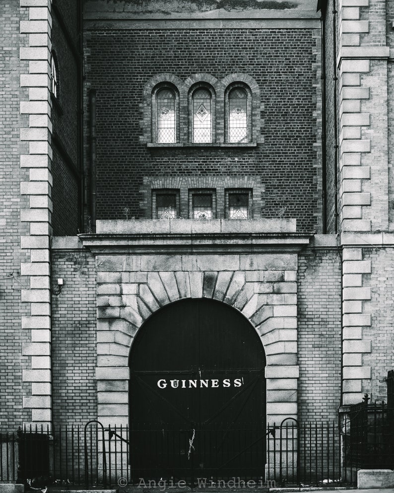 Puede incluir: Una foto en blanco y negro de un edificio de ladrillo con una gran puerta arqueada. La puerta es de metal negro con la palabra "GUINNESS" escrita en letras blancas.