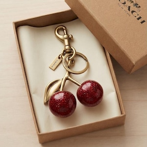 May include: A Coach brand cherry-shaped keyring featuring two round, glittery red spheres attached to a brass-toned metal stem with leaves, a split ring, and a lobster clasp, presented inside a brown cardboard box with the Coach New York logo.