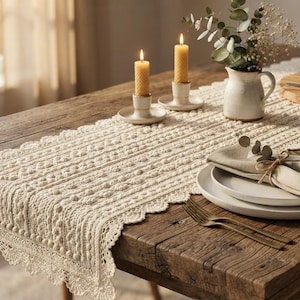 May include: A cream-colored crocheted table runner with a textured bobble stitch pattern and a scalloped lace edge. The runner is laid out on a wooden table, featuring two lit beeswax candles in holders, a small ceramic pitcher, and plates.