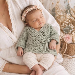 Può includere: Un maglione a portafoglio a maniche lunghe in uncinetto verde salvia con chiusura a fiocco laterale, abbinato a pantaloni in maglia a coste color crema. Il neonato indossa una delicata fascia floreale con piccoli fiori rosa e bianchi.