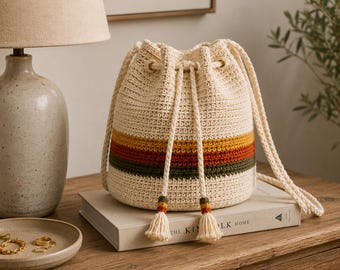 Puede incluir: Un bolso tipo cubo de ganchillo color crema con rayas horizontales en amarillo mostaza, naranja quemado y verde oliva. El bolso tiene un cierre de cordón con dos cuerdas trenzadas largas que terminan en borlas a juego con detalles.