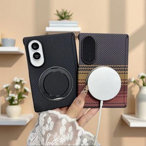 Può includere: Due cover rettangolari per telefono con aperture per la fotocamera. La cover sinistra è nera con una trama in fibra di carbonio e un anello magnetico circolare. La destra presenta un motivo a griglia scuro con sfumatura orizzontale.