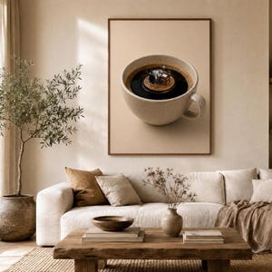 A framed wall art print featuring a surreal image of a miniature office worker sitting at a desk inside a cup of coffee, hanging above a beige sofa in a brightly lit living room.