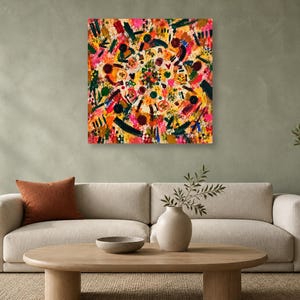 A square abstract colorful painting hangs on a green wall above a beige sofa with an orange pillow, next to a wooden coffee table and an olive tree in a pot.