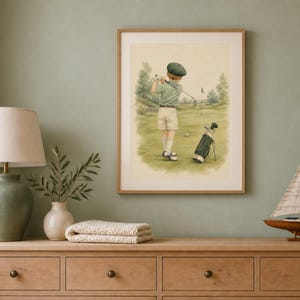 May include: A framed wall art print depicting a person in a green shirt, beige shorts, and a cap swinging a golf club on a green course. A golf bag stands nearby on the grass. The artwork is displayed in a light wood frame on a sage green wall.