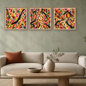A living room interior featuring a beige sofa with an orange pillow, a wooden coffee table, and three square framed abstract prints on the wall.