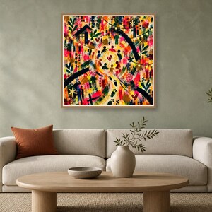 A framed square abstract colorful print mounted on a greenish wall above a beige sofa and coffee table.